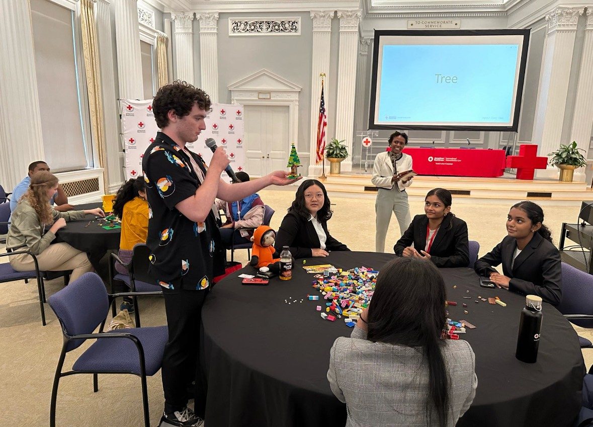 someone standing and speaking on a microphone in a room full of other youth action campaign members sitting at round tables with an American Red Cross table in the background