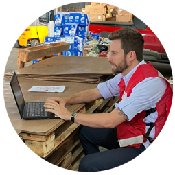 A Red Cross volunteer in a warehouse full of supplies working on his computer