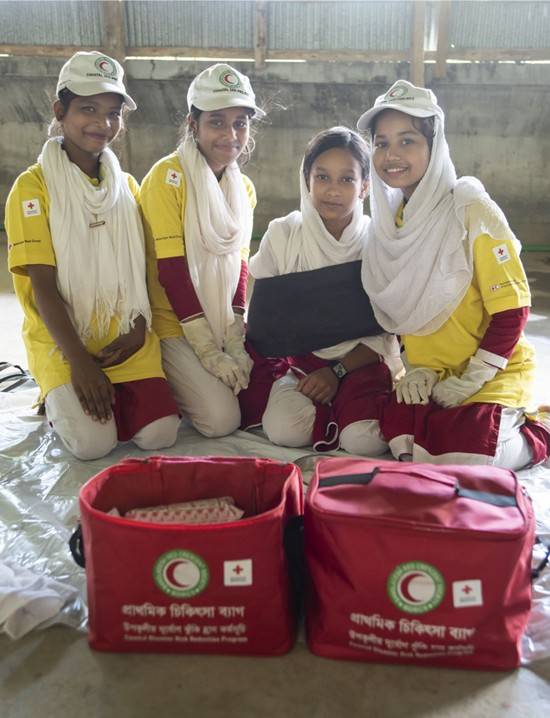 4 girls are kneeling down behind a variety of disaster relief supplies.