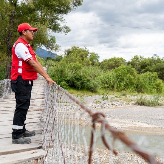A Red Cross volunteer is standing on a bridge observing a flooded river