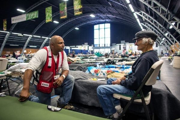 Red Cross volunteer speaking with man in a shelter full of cots.