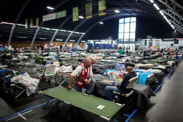 Red Cross volunteer speaking with man in room full of cots.