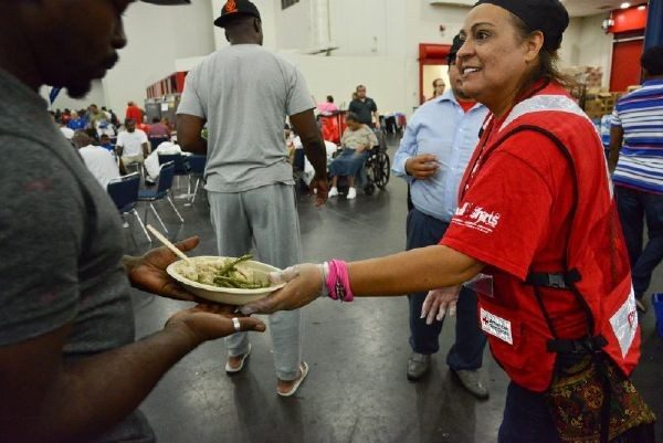 Red Cross volunteer handing plate of food to man with group of people in the background.