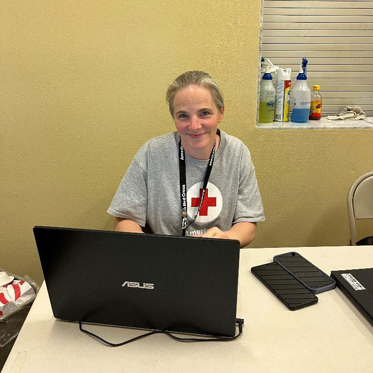 Angella Working sitting at desk, working on laptop and wearing a Red Cross t-shirt.