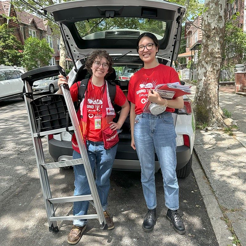 Hannah and Kehui standing on street with ladder and gear to install smoke alarms.