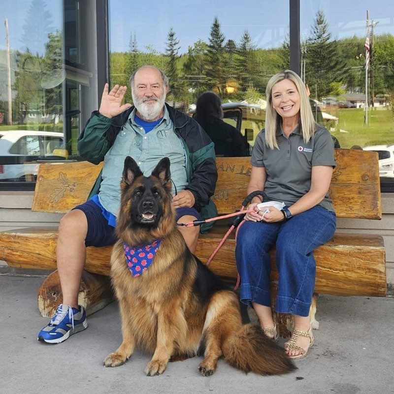 Two people sitting on a wood bench with a service dog sitting and wearing a scarf around it's neck.