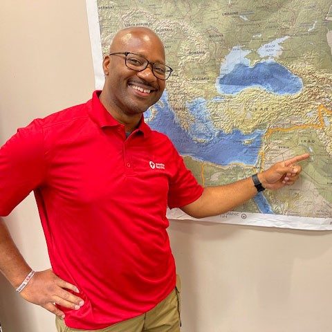 Nasir Ahmad wearing glasses and an American Red Cross shirt, points at a map that's hanging on the wall.