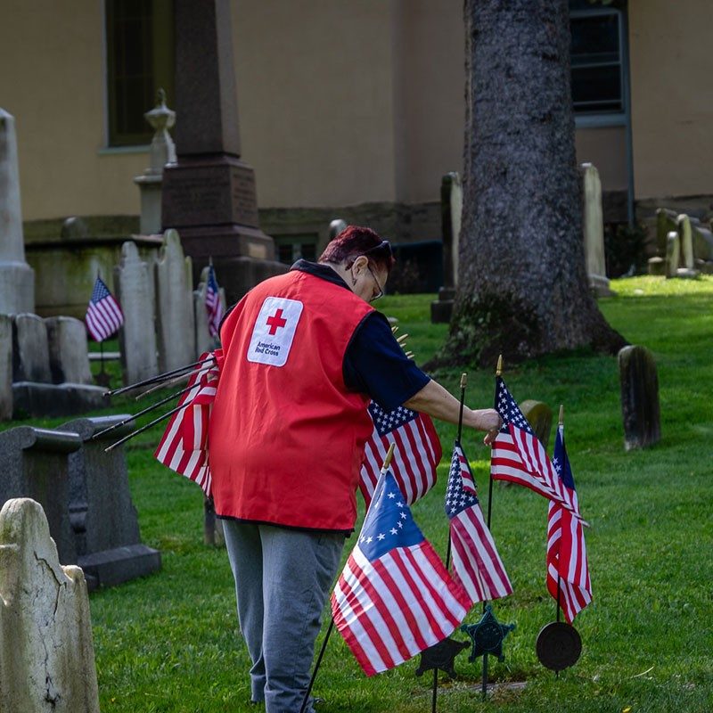 American Red Cross volunteer putting small American flags next to headstones in cemetery.