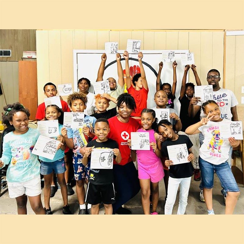 classroom of children holding paper up with a Red Cross worker in the middle of them.