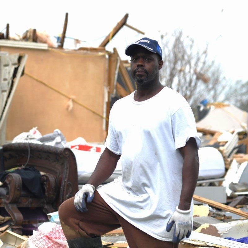 Carl Duncan, wearing work gloves, standing in the middle of debris by a damaged house.
