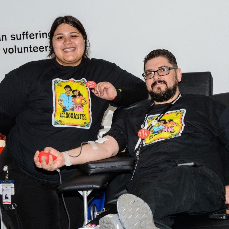 A person giving blood in a Los Danantes shirt with another person satnding next to them.