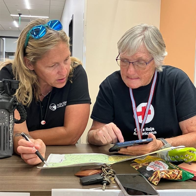 Two red cross volunteers sitting at desk with papers and talking to each other.