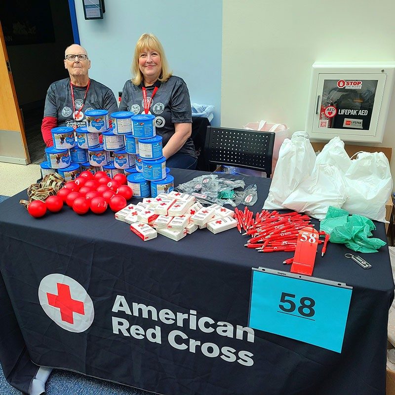 Two Red Cross volunteers sitting at a table with Red Cross stress balls, pens, tissues, keychains and other items.