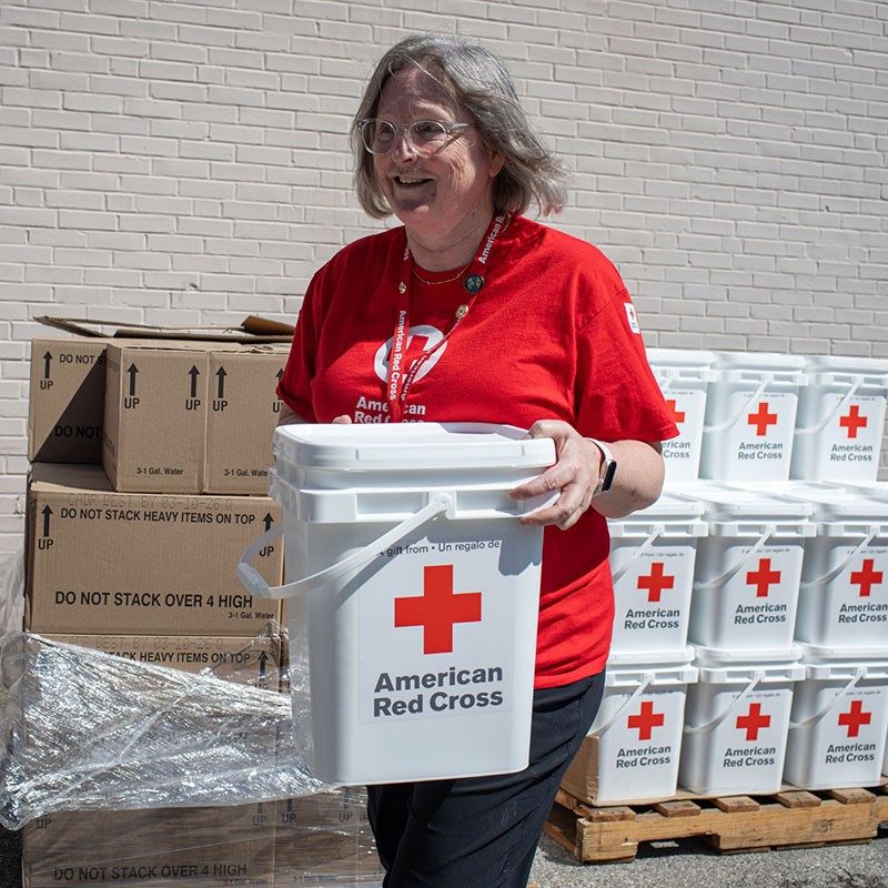 Red Cross volunteer carrying a 5 gallon Red Cross bucket, with a pallet of Red Cross 5 gallon buckets in the background.