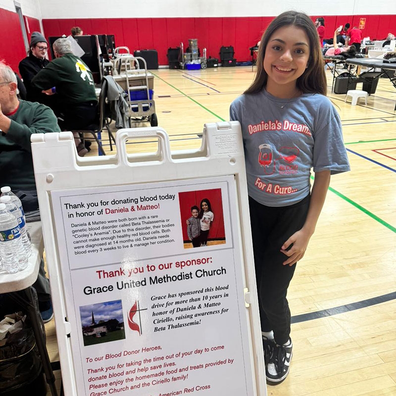 Red Cross volunteer Daniela at a blood donation event standing next to a Red Cross blood donation event sign.