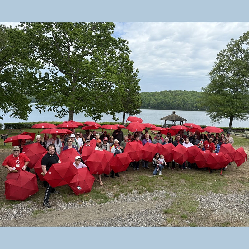 large group pic of Red Cross volunteers all holding red umbrellas by a lake. 