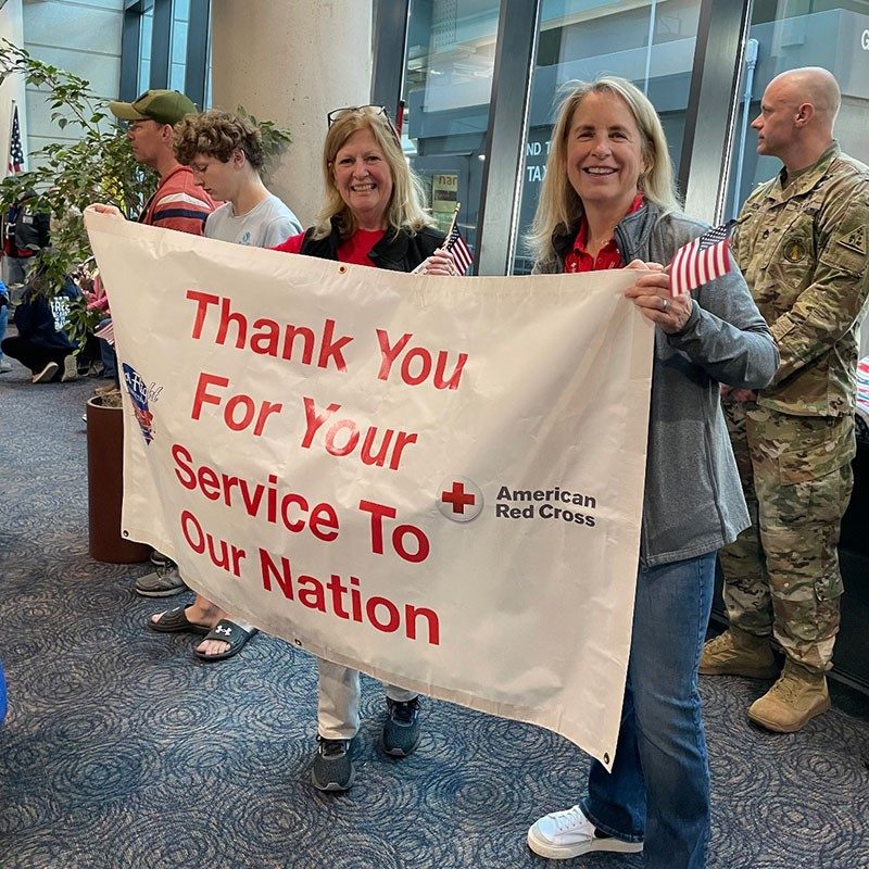 Red Cross volunteers standing in an airport lobby and holding a large banner that says, "Thank you for your service to our nation".