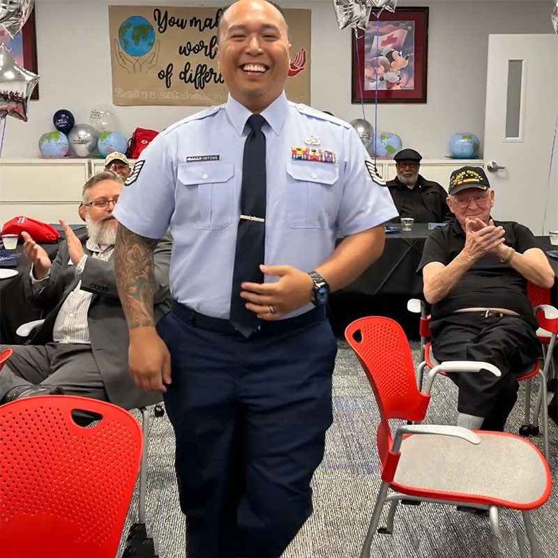 Military member in military dress clothing, in an office with other people sitting and clapping.