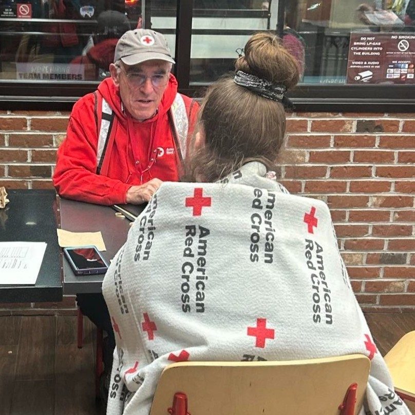 Red Cross volunteer helping a disaster victim who is wrapped in a Red Cross blanket.