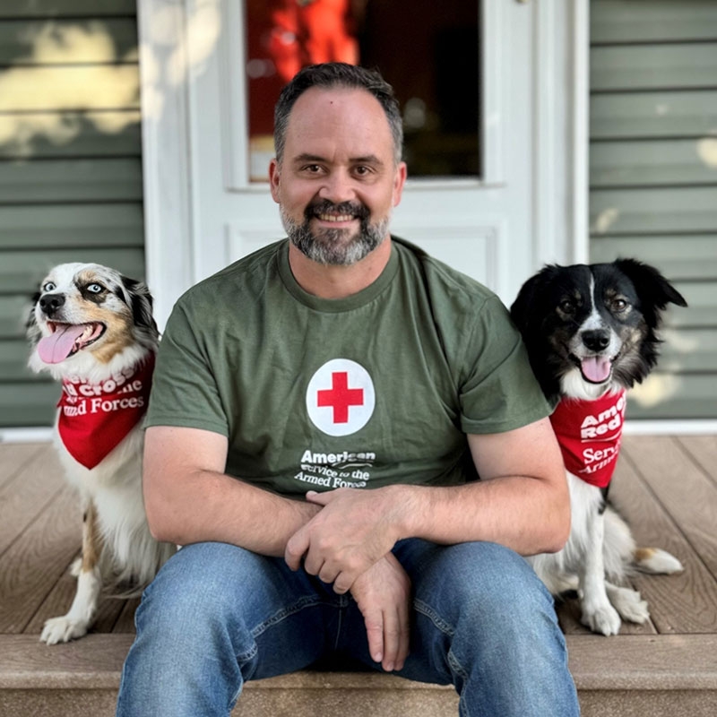 Logan, wearing a Red Cross shirt, and therapy dogs Nova and Lucy, wearing Red Cross scarfs, sitting on a porch.