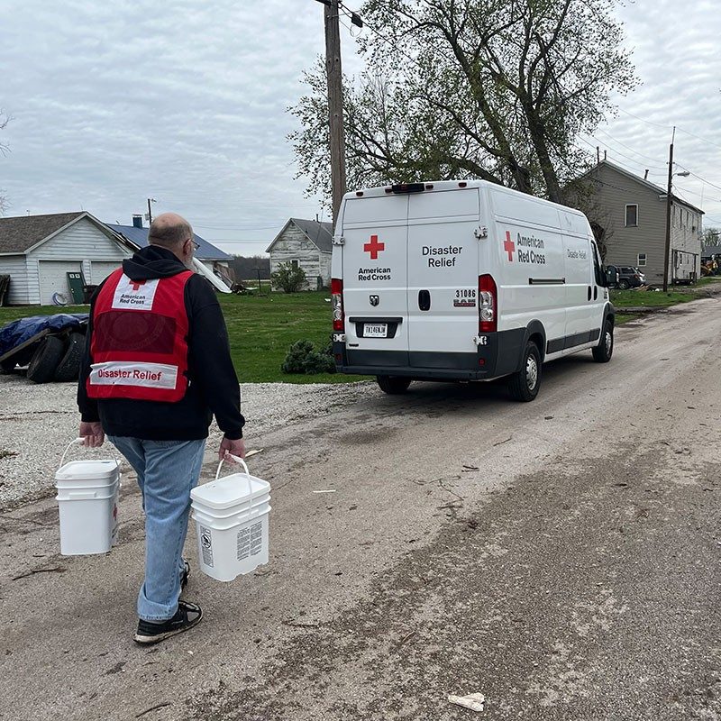 Red Cross volunteer carrying two 5 gallon buckets walking down a dirt road towards a Red Cross van.