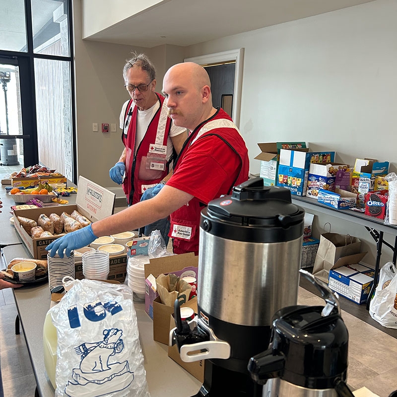 Two Red Cross volunteers serving coffee and doughnuts that are spread out on tables.