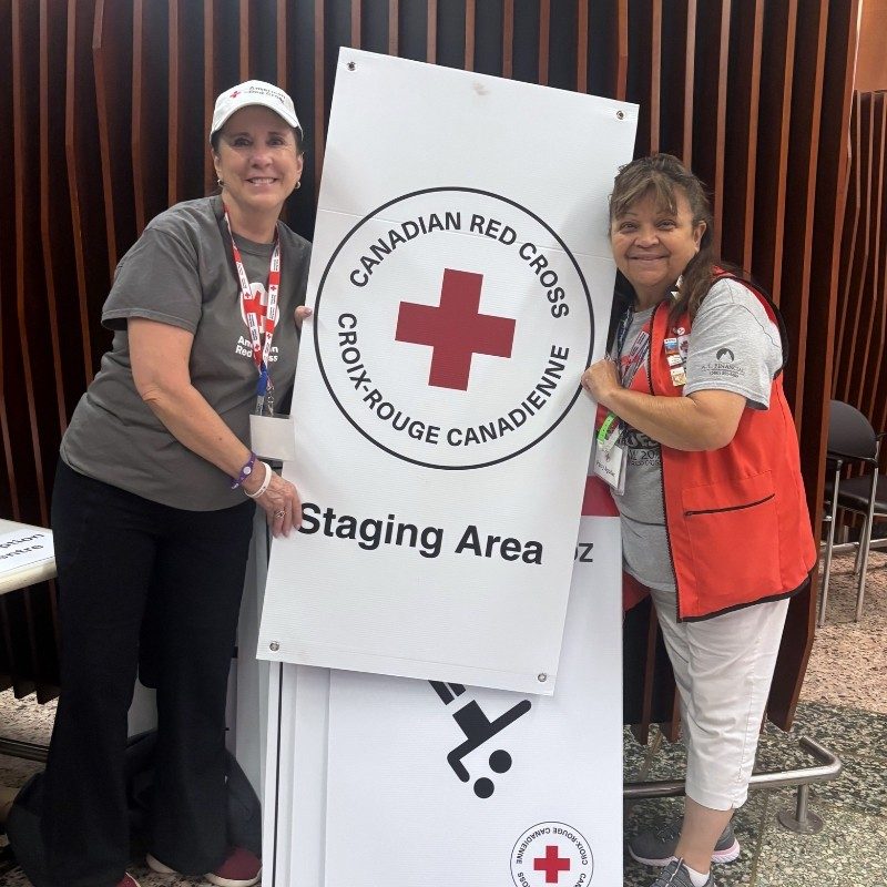 Two Red Cross volunteers standing on each side a "Canadian Red Cross" poster.