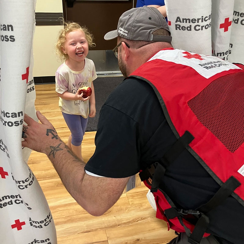 Red Cross volunteer making a small child, who's holding a bitten apple, laugh.