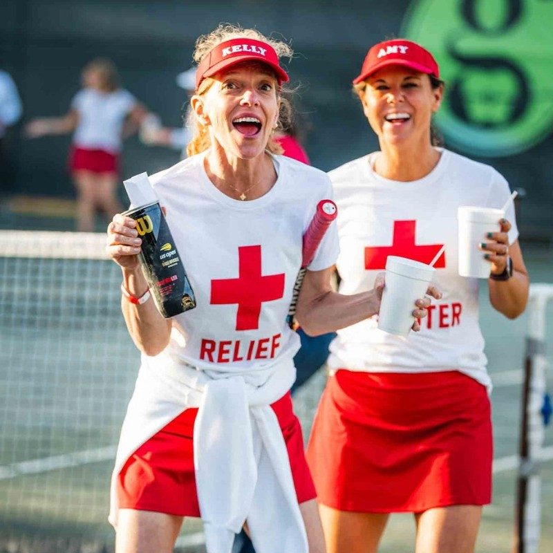 Two tennis players on tennis court wearing Red Cross shirts.