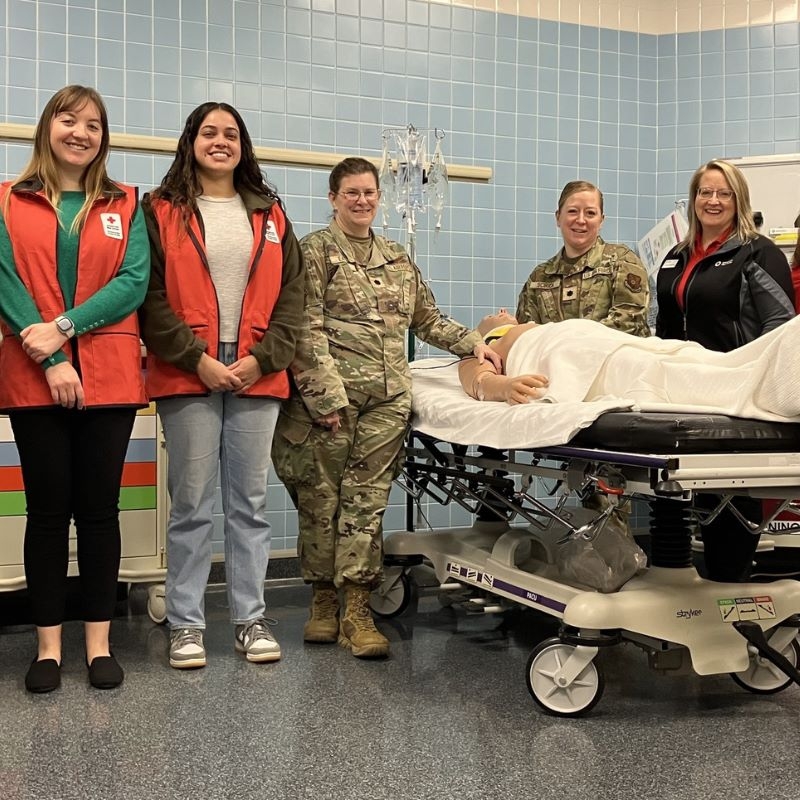 Three Red CRoss volunteers and three military memebers standing next to medical mannequin on a bed.