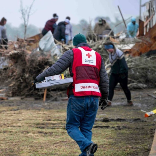 Red Cross Volunteer, carrying emergency supplies and walking in front of a tornado damaged house.