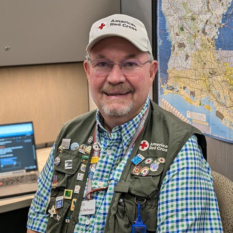 John Mathews sitting at a desk and wearing a red cross vest and hat with multile buttons and patches on his vest.