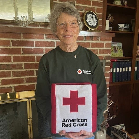 Pearl Gruenke standing next to a fireplace in her home and holding a Red Cross cloth bag.