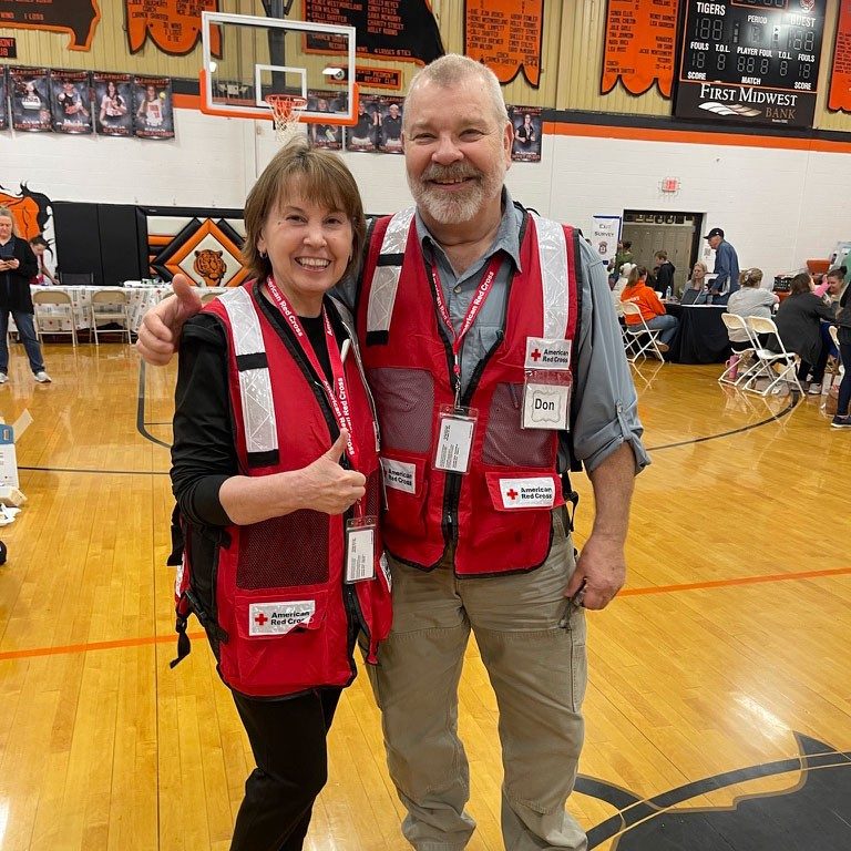 Red Cross volunteers Pasty and Don Berry wearing Red Cross vests while standing in a Red Cross shelter.