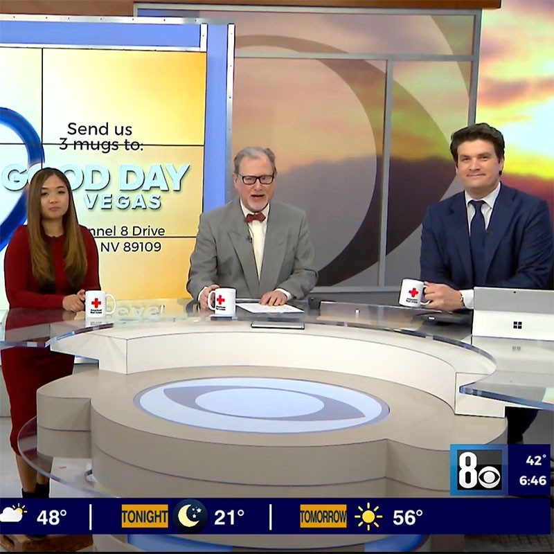 three newscasters holding Red Cross coffee mugs while sitting at a desk during the news broadcast.