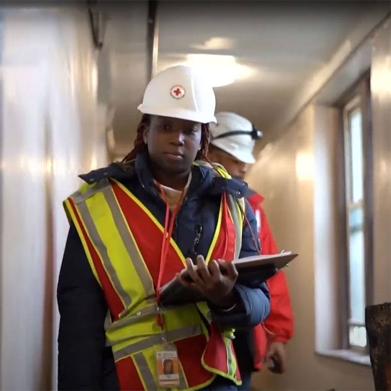 Red Cross volunteer wearing hard hat, vest, and holding a clipboard.