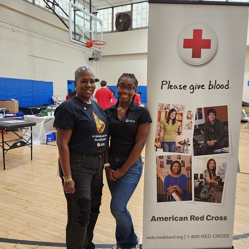 Taylor Williams and another person standing next to a Red Cross please give blood banner in a gym.