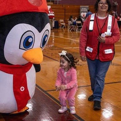 Young child standing near a penguin dress-up character and a Red Cross volunteer.