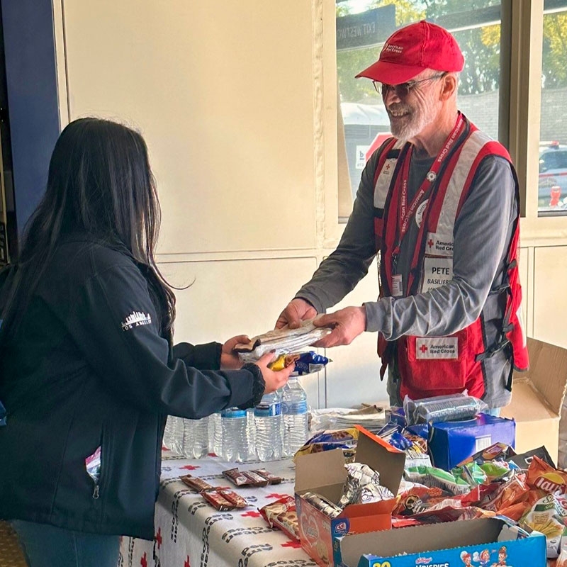 Red Cross volunteer, standing next to a table with food and drinks, hands items to a local resident in need.