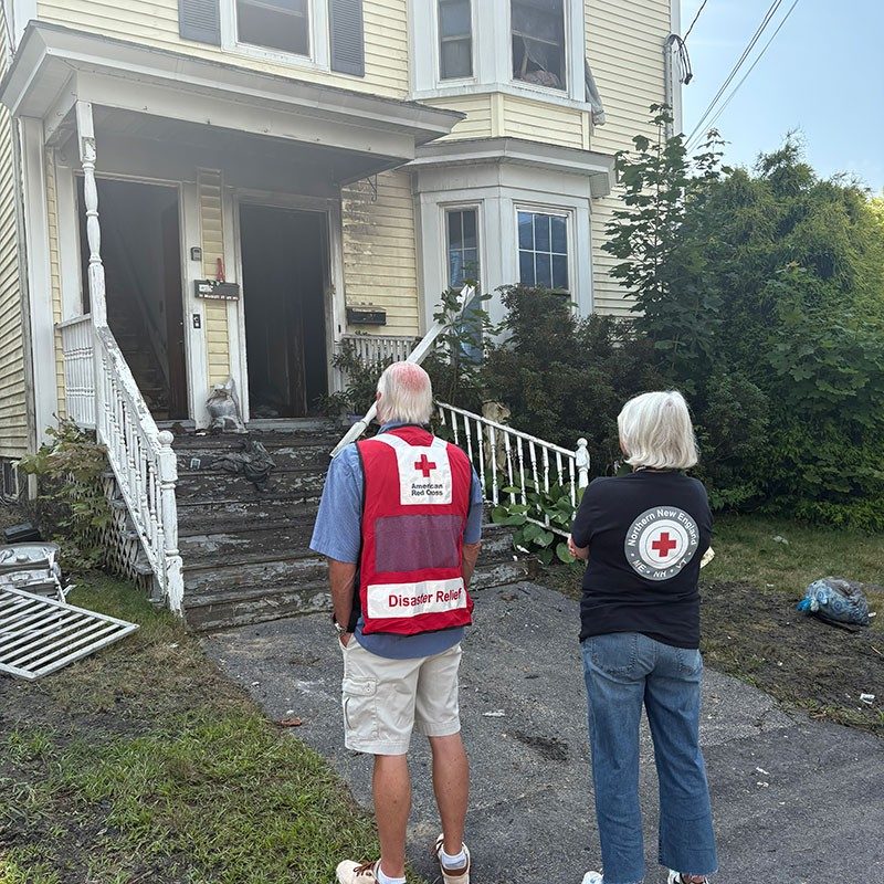 Two Red Cross volunteers standing in front of a burned house.