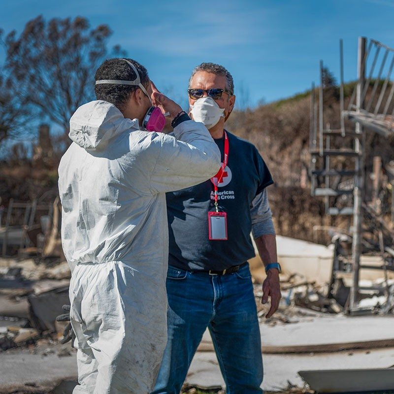 Red Cross volunteer, wearing a mask, standing next to person wearing a mask and protective clothing with debris in the background.