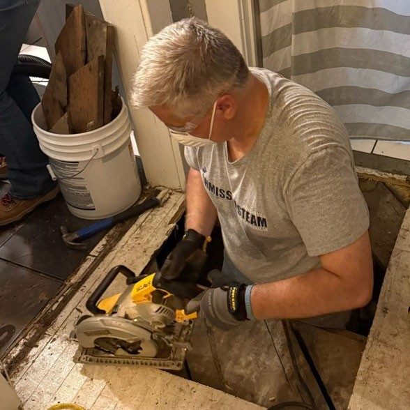 person wearing mask and using a skill saw, cuts a hole in a wooden floor.