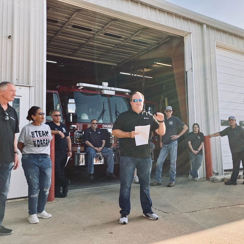 Group of people standing in front of a fire truck at a fire house.