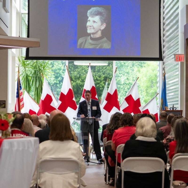 Person standing at a podiumspeaking to a croward with Red Cross flags behind him.