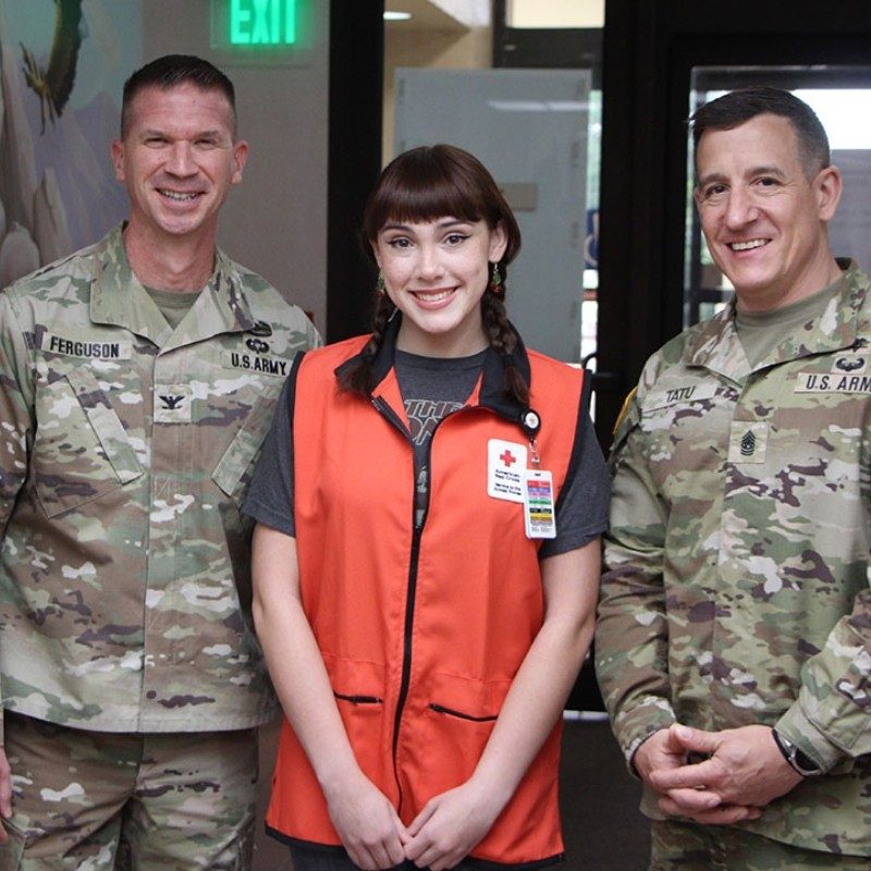 Two service members in uniform standing on each side of a Red Crossteen volunteer.