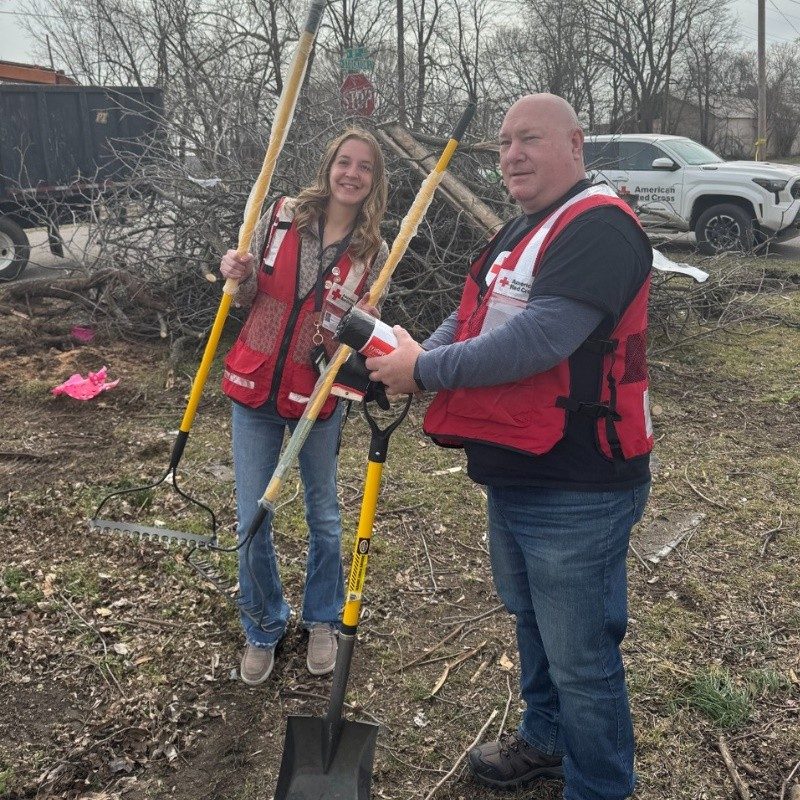 Two Red Cross volunteers in vest holding shovels in hand at a disaster site.