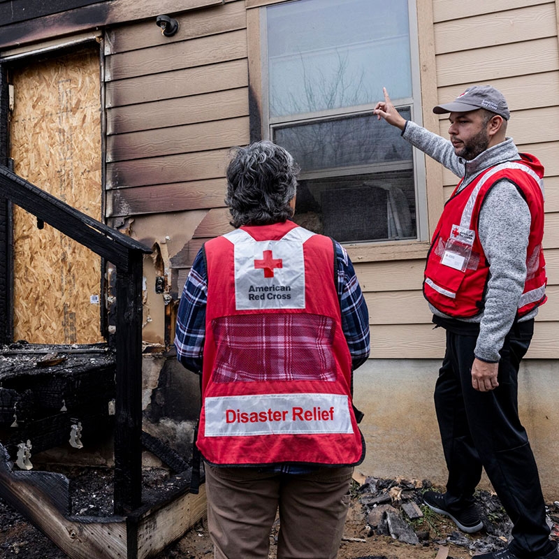 Two Red Cross volunteers talking with each other next to a house that has fire damage.