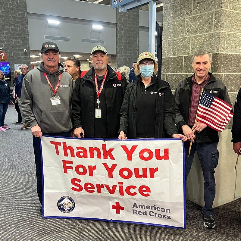 Red Cross volunteers standing in an airport lobby and holding a large banner that says, "Thank you for your service".
