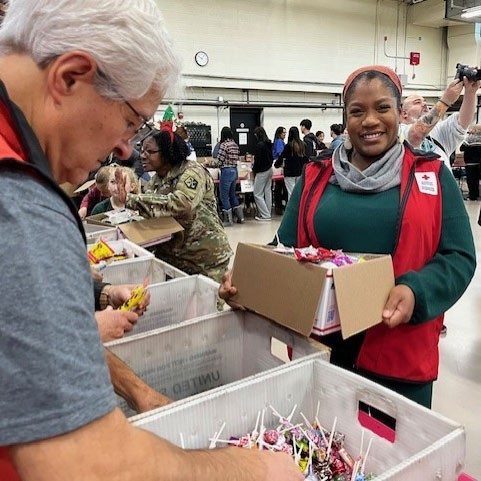 Red Cross volunteer, in a room with other volunteers, guests, and tables full of candy, loads a box with holiday candy.