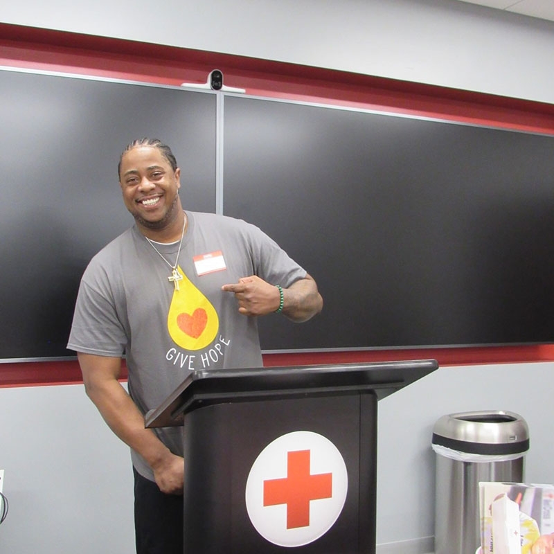 man wearing and pointing at his give hope blood donor shirt, stands at a podium with a Red Cross logo on it.